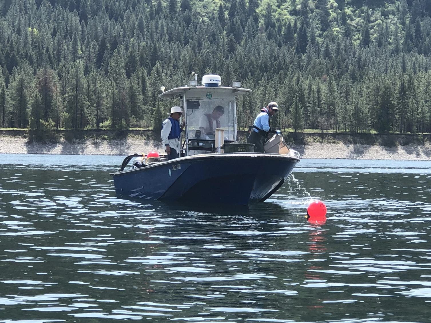 Three people wearing life jackets work from a small blue research boat on a calm lake. One person stands near the bow pouring water from a container beside a bright red buoy floating in the water. Forested hills line the shoreline in the background.